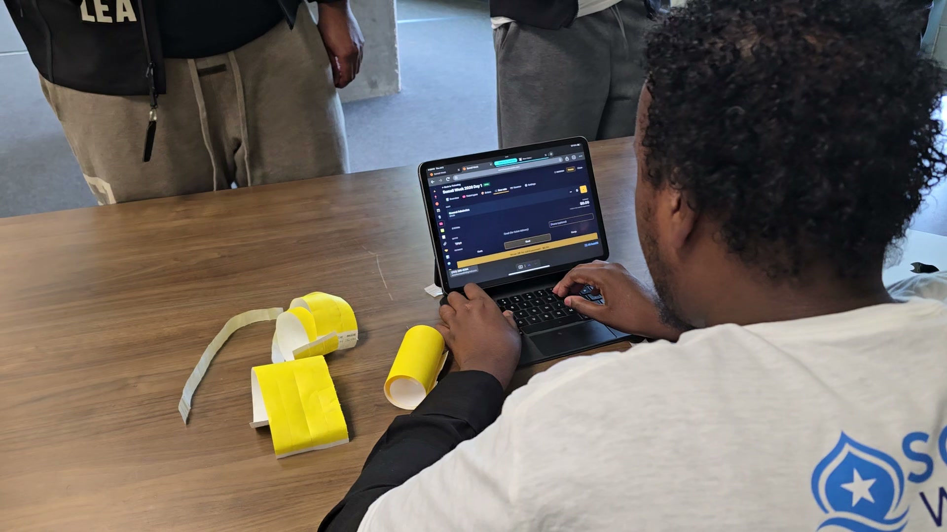 A Somali Week 2026 volunteer running door ticket sales on a laptop with the ScoreLM admin open — yellow wristbands on the table next to the keyboard